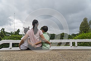 Back view of loving Asian mother hug her kids sitting on bridge, caring black mom embrace child, relaxing looking to black cloud,