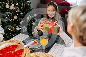 Back view of loving grandmother giving festive box with Christmas present to adorable granddaughter sitting at dinner