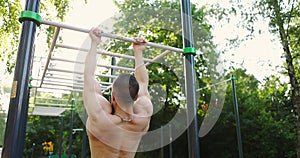 Back view of male athlete doing pull-up on horizontal bar outdoors while using street gym equipment