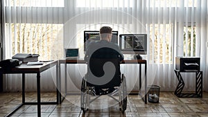Back view of male web developer in a wheelchair writing program code on multiple computer screens while sitting at his photo