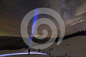 Back view of man with head flashlight standing on snowy valley under beautiful dark blue winter starry sky, bright blue beam on