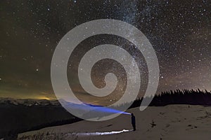 Back view of man with head flashlight standing on snowy valley under beautiful dark blue winter starry sky, bright blue beam on