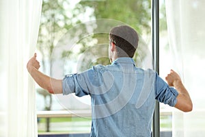 Back view of man opening curtains and looking away