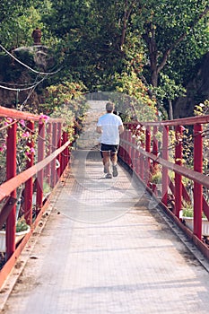 Back view Man run across red iron suspension bridge way to forest park. Outdoor Sport after quarantine, explore new life
