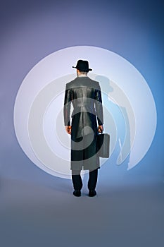 Back view of man silhouette in black coat and hat holding briefcase in the spotlight on studio background
