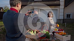 Back view man taking photo of woman posing with organic local products on farm market. Shooting over shoulder of loving