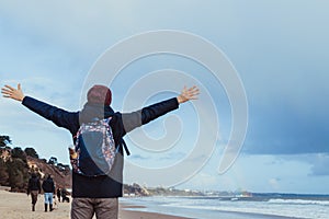 Back View man in warm clothes raised arms up to the sky, enjoying rainbow view above the sea during a walk on winter