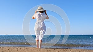 back view on a mature woman in a straw hat and white dress walks along the blue sea coast on a sunny summer day