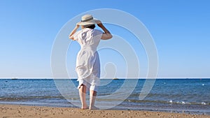 back view on a mature woman in a straw hat and white dress walks along the blue sea coast on a sunny summer day