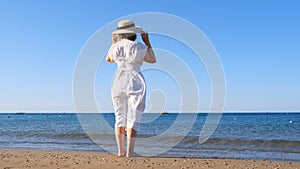 back view on a mature woman in a straw hat and white dress walks along the blue sea coast on a sunny summer day