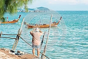 Back view of a mid aged man tourist in casual dress taking photo of local long tail boat with smartphone in front sea of Phuket