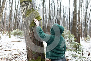 Back view of middle-aged woman standing, touching green moss on trunk of tree with hand in park forest in snowy winter.
