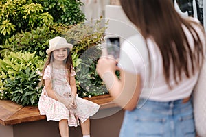 Back view of mom takes photos of her daughter on her phone. Adorable little girl sitting on bench. Focus on kid