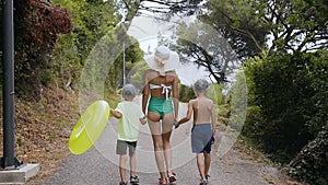 Back view. Mom and two sons are walking in the park by the sea. A young woman in a swimsuit and a big hat along with