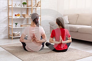 Back view of mother and daughter sitting in lotus pose, making namaste gesture, doing morning yoga practice together