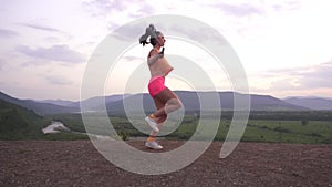 Back view on muscular young brunette woman jumping with skipping rope on the mountain peak. Green mountain ladscape