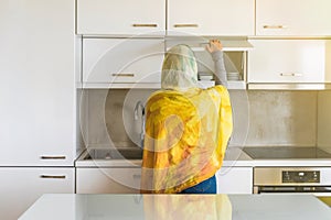 Back view on a muslim woman in a scarf opens door of a whte cabinet with cups and plates.