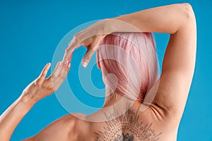 Back view of naked tattooed woman with pink hair and perfect skin posing with raised hands isolated over blue studio