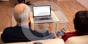 Back view of old couple in their living room looking a laptop with white isolated mock-up