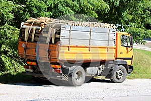 Back view of old yellow truck filled with firewood parked on gravel parking lot next to paved road surrounded with dense trees