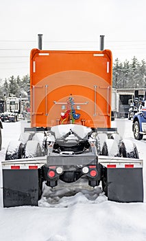 Back view of orange big rig semi truck with Fifth wheel standing on covered with snow winter parking lot