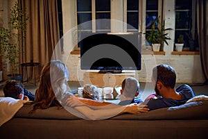 Back view of parents and children spending evening together, sitting on couch in living room and watching tv, eating