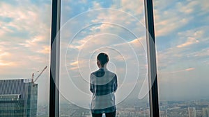 Back view of pensive woman looking at cityscape through window of skyscraper