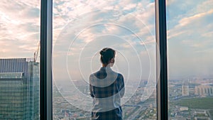 Back view of pensive woman looking at cityscape through window of skyscraper