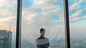 Back view of pensive woman looking at cityscape through window of skyscraper