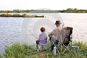 Back view portrait of adult man and teenage boy sitting together on lake fishing with rods