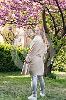 Back view portrait of blonde girl against background of sakura blossoms tree. Young beautiful woman walking in spring blooming