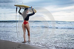 back view portrait of young happy surfer girl in red swimsuit walking towards the sea carrying yellow surf board and ready