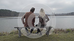 Back view of positive man and woman sitting on bench in front of forest lake and talking. Wide shot of happy loving