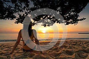Back view of pregnant woman sit on sand and watching sunset at tropical beach. Woman wear swimsuit and straw hat relaxing