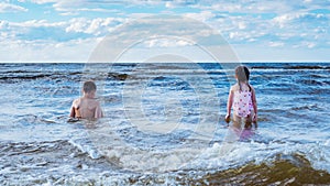 Back view of preteen boy brother, little girl sister siblings family sitting playing in water on beach near foamy waves.