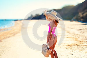 Back view of pretty girl in straw hat and bikini walks sea of white sand at sunny day holds handbag