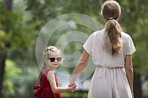 Back view of pretty small blond long-haired child girl in sunglasses and fashionable red dress holding trustingly hand of young