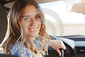 Back view of pretty smiling woman with broad smile, has attractive look, sits at wheel in car, has break after long trip, looks di