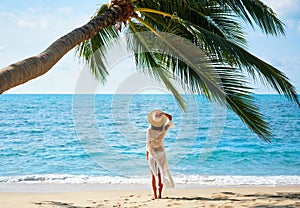Back view of pretty young woman relax and enjoy sea standing under palm tree on tropical beach
