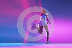 Back view. Professional female athlete running away isolated on blue studio background in mixed pink neon light. Healthy