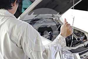 Back view of professional young mechanic man in uniform holding wrench against car in open hood at the repair garage.