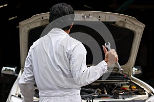 Back view of professional young mechanic man in uniform holding wrench against car in open hood at the repair garage.