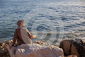 Back view. relaxed girl in a hat and coat sitting in front of the sea on the stones