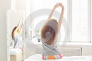 Back view of relaxed woman stretches in bed, poses near window against cozy bedroom interior, wakes up in morning after good sleep