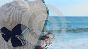 Back view of sad young woman in hat sitting alone on beach, looking at sea waves