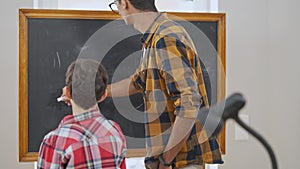 Back view of schoolboy standing at chalkboard with teacher writing numbers in slow motion. Interested Middle Eastern