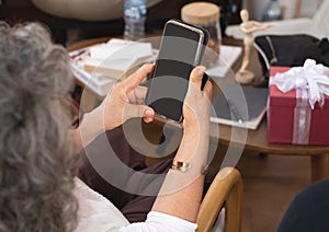 Back view of senior elder asian grandmother holding smart phone tablet sit on sofa or couch in living room at home