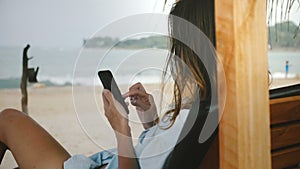 Back view shot of young woman using smartphone shopping app relaxing in lounge beach chair with amazing seaside scenery.