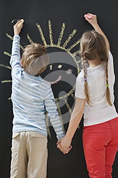 Back view of siblings drawing sun on blackboard while holding hands