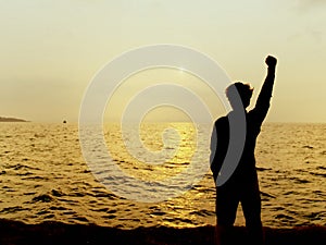 Back view of silhouette of man raising hands during sunset over the sea. Successful concept.